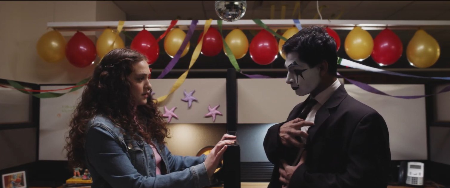 Still from Fools Rush In: a woman and a man in mime makeup talk in a cubicle decorated with balloons